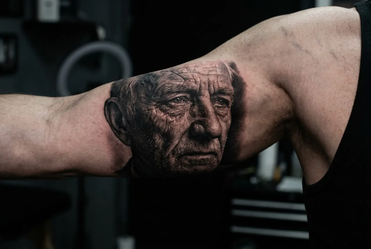 A black-and-grey realism portrait of an elderly man's weathered face on an upper arm.