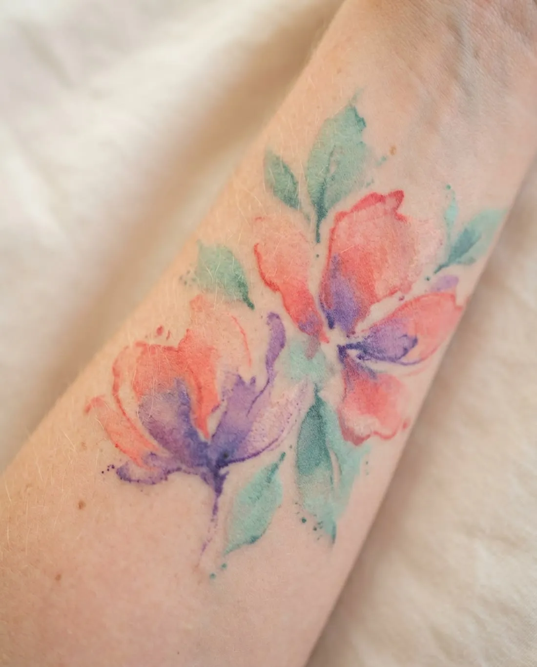 Healed watercolor peony tattoo in coral and violet on a forearm